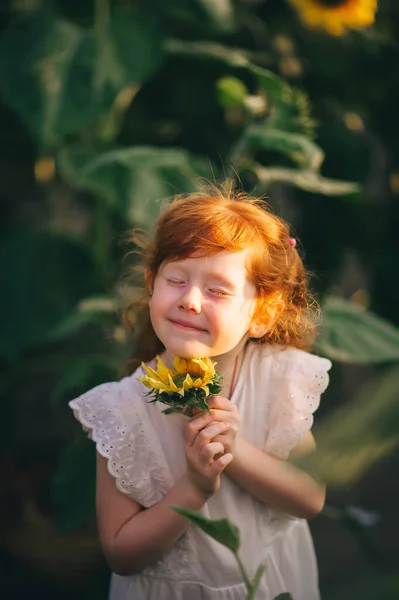 Child in a field of sunflowers. The little redhead girl closed her eyes and make a wish with closed eyes. Closeup portrait