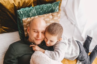 Mutlu kel baba ve oğlu rahatlıyor, yatakta birlikte uzanıyor, aile eğleniyor, sarılıyor ve sarılıyor, gülümsüyor baba ve çocuk hafta sonunu evde geçiriyor. Sıcak bir ilişki. Yakın çekim..