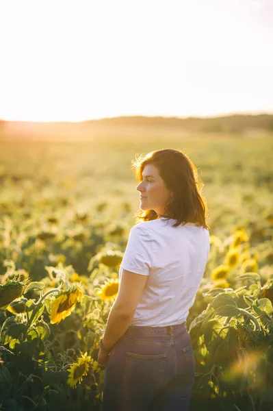 Beautiful young woman enjoying nature on the field of sunflowers at sunset. Caucasian woman in a white shirt and blue jeans enjoys summer and vacation.