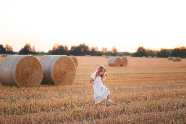 穿着白色衣服的可爱的小女孩在落日的时候带着一大捆麦子漫步在麦田里 夏天户外麦田里可爱的小女孩 — 图库照片