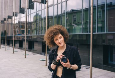 Ceketli, gülümseyen kıvırcık kadın modern binaya karşı şehir caddesinde poz veriyor. Geleneksel yapı mermerden yapılır.