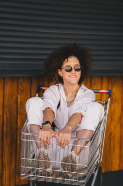 Afro saçlı ve güneş gözlüklü havalı bir kız alışveriş arabasında otururken eğleniyor. Yakın plan portre