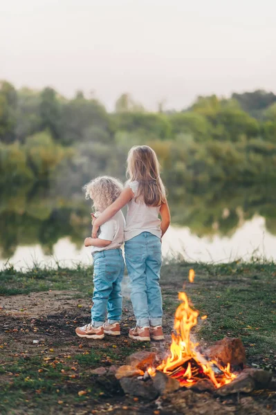 Two cute little girls are standing by the fire and watching the sunset with their backs. Family leisure with kids at summer.