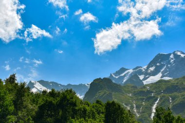 Kafkas Dağları 'nın karla kaplı zirvelerinin güzel bir manzarası. Mavi gökyüzüne karşı beyaz bulutlar.