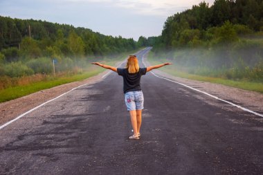 Genç, kızıl saçlı bir kadın, bir yaz akşamı uzaktaki yola bakıyor. Yol, ağaçlar, çimenler olan kırsal alan. Arka planda sis var.