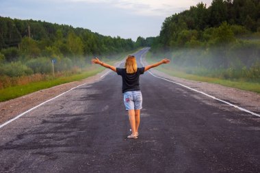 Genç, kızıl saçlı bir kadın, bir yaz akşamı uzaktaki yola bakıyor. Yol, ağaçlar, çimenler olan kırsal alan. Arka planda sis var.