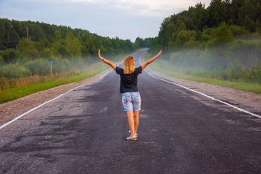 Genç, kızıl saçlı bir kadın, bir yaz akşamı uzaktaki yola bakıyor. Yol, ağaçlar, çimenler olan kırsal alan. Arka planda sis var.