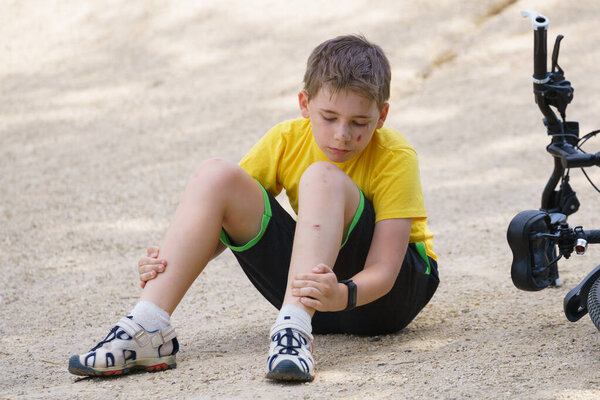 Мальчик с поцарапанным лицом сидит на земле и исследует свои раны на ноге
.