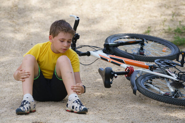 Мальчик с поцарапанным лицом, упавший с велосипеда, садится на землю и исследует свои раны на ноге
.