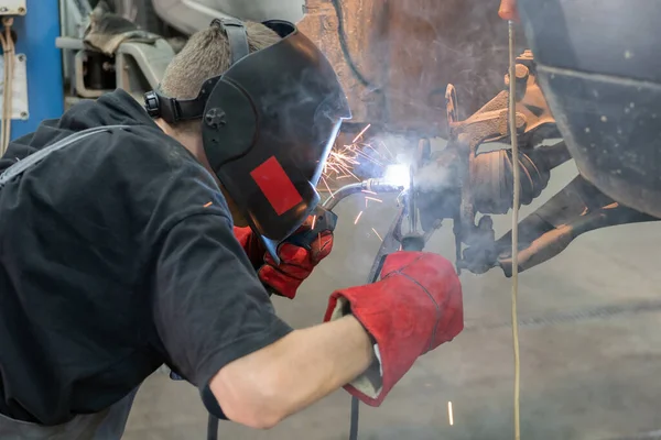 A car mechanic in a welding mask welds broken off wheel bolts to the car hub