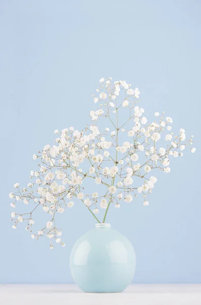 Light soft elegant home decor with small airy flowers in glossy pastel blue vase on wood table and blue wall, vertical.