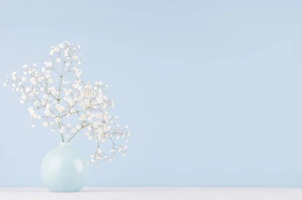 Light soft elegant home decor with small airy flowers in glossy pastel blue vase on  wood table and blue wall.