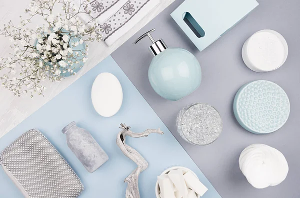 Fashion cosmetic products set on pastel blue, grey color paper - white soap, towel, flowers, soap dispenser, blue ceramic vase, silver cosmetic bag, top view.