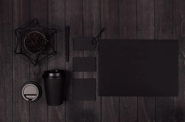 Coffee takeaway set mockup for brand - black paper cup, blank paper, card, label, decoration, coffee beans on dark black wood board, top view.