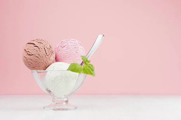 Sweet ice cream balls - strawberry, chocolate, creamy in elegant transparent glass bowl with spoon, green mint on white wood table, pink wall