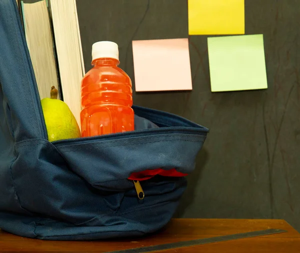 juice and pear in a backpack with textbooks on the desk against the background of the school board with stickers. school concept, education, lunch break.