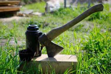 Tahta kirişe sıkışmış bir baltanın yanında bir termos ve bir fincan sıcak çay. Turizm ve açık hava eğlencesi kavramı