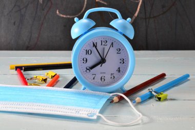 Okul malzemelerinin arka planında mavi tıbbi maske ve masada bir çalar saat..