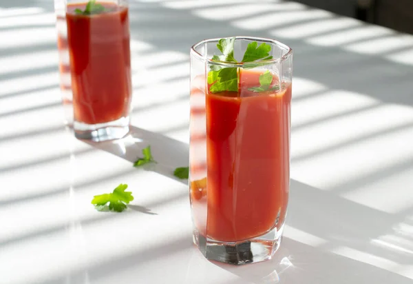 red tomato juice in glass glasses with parsley leaves. the shadow of the blinds on the windows falls on the table. copy space.