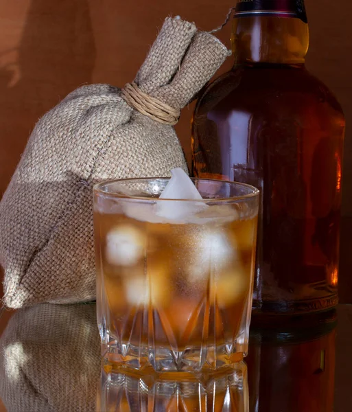 Bicchiere misted di whisky con ghiaccio e bottiglia con riflessione su fondo iuta — Foto Stock