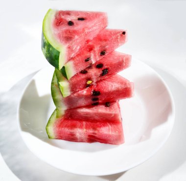 Arka plandaki üçgen karpuz parçaları beyaz bir masanın arka planındaki beyaz bir tabakta, üst tarafta. Hasat konsepti, vitaminler lezzetli ve sağlıklı yiyecekler. Boşluğu kopyala.