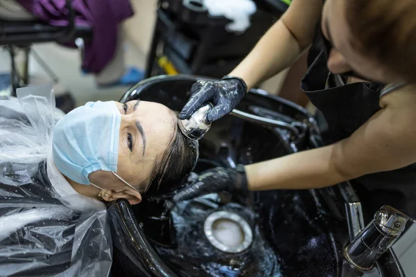 Hairdressing services during the coronavirus. Hands of hairdresser washing head of woman in mask. Hairdresser washing head of woman