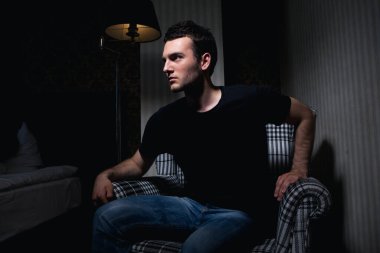 Siyah tişörtlü yakışıklı esmer adam gölgelerdeki kareli sandalyede otururken meydan okuyor.