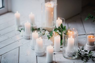 Cam şamdanların içindeki beyaz mumların güzel dekoru yeşil yaprakların ve çiçeklerin arasında beyaz ahşap bir zeminde duruyor.