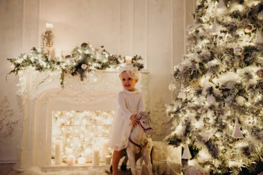 Beyaz elbiseli sevimli çocuk Noel ağacı ve şömineli at oyuncağının üstünde.