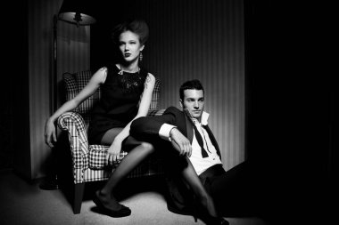 Odadaki gölgelerde klasik giysiler içinde tutkulu güzel bir çift: sandalyede oturan kadın, ayaklarının dibinde yatan bir adam.