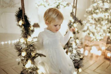 Noel stüdyosunda salıncakta sallanan beyaz elbiseli sevimli çocuk.