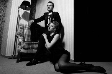 Odadaki gölgelerde klasik giysiler içinde tutkulu güzel bir çift: sandalyede oturan adam, ayaklarının dibinde yatan kadın.