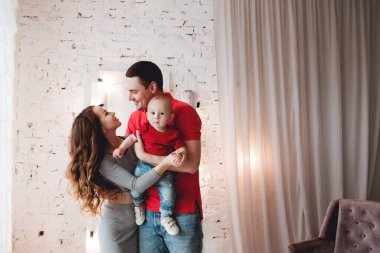 Anne ve baba, ellerinde aynı kotlu ve kırmızı tişörtlü küçük bir çocuk, parlak bir odada, perdeleri, merdivenleri ve ampulleri olan beyaz tuğladan bir duvara karşı duruyor.