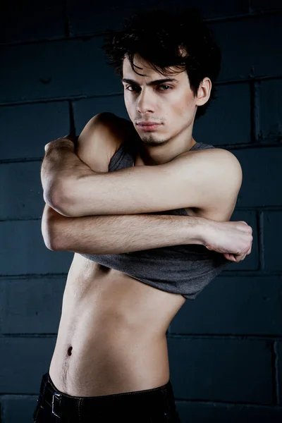 Genç, zayıf, koyu saçlı bir adam gri tişörtünü çıkarıyor.
