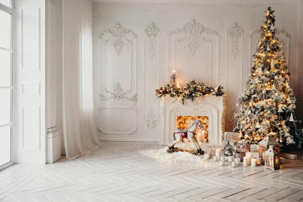 Beyaz bir odanın içinde Noel ağacı, süslü bir şömine, sallanan at ve yeni yıl hediyeleri olan bir oda.
