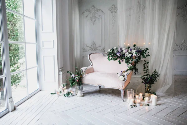 花や緑で装飾された柔らかいピンク色のヴィンテージソファは 大きな窓やカーテンの近くのガラスの燭台のろうそくに囲まれた白い木製の床の古典的な部屋に立っています — ストック写真