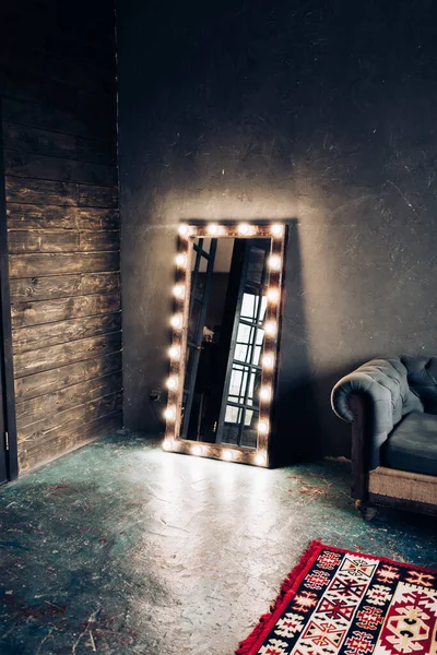 Corner room with a wooden and gray cement wall, an aged floor, black glass doors and electric dressing makeup mirror with lights bulbs and oxford sofa in loft style