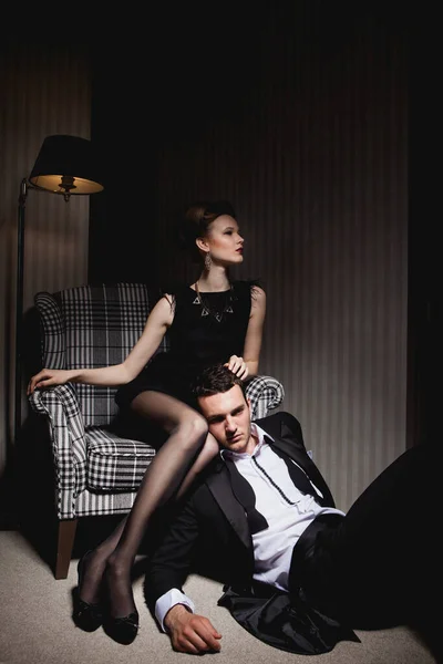在房间的阴影中 穿着经典衣服的年轻貌美的情侣们 坐在椅子上的女人 在她脚边的男人 图库照片