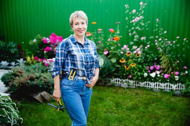 Kot pantolonlu yaşlı bir kadın ve kareli bir gömlek. Arka planda güzel çiçek bahçesinin budama makinesini tutuyor.