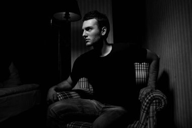 Siyah tişörtlü yakışıklı esmer adam gölgelerdeki kareli sandalyede otururken meydan okuyor.