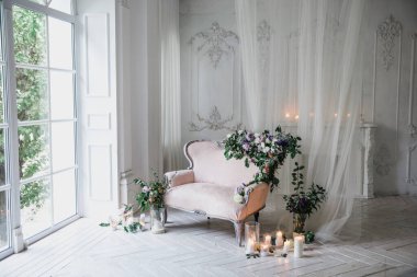 Çiçekler ve yeşillerle süslenmiş, yumuşak pembe renkli kanepe, büyük pencere ve perdelerin yanındaki cam şamdanlarla çevrili beyaz ahşap bir zeminde klasik bir odada duruyor.