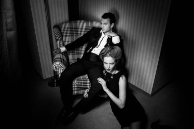 Odadaki gölgelerde klasik giysiler içinde tutkulu güzel bir çift: sandalyede oturan adam, ayaklarının dibinde yatan kadın.