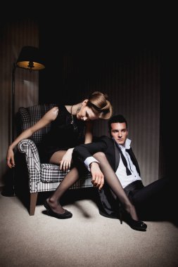 Odadaki gölgelerde klasik giysiler içinde tutkulu güzel bir çift: sandalyede oturan kadın, ayaklarının dibinde yatan bir adam.