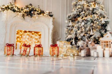 Kırmızı-turuncu oyuncak tren, beyaz zeminde ışıkları olan bir Noel ağacının arka planında ışıklar, çelenkler ve şöminesi olan.