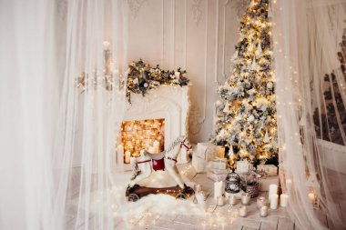 Beyaz bir odanın içinde Noel ağacı, süslü bir şömine, sallanan at ve yeni yıl hediyeleri olan bir oda.