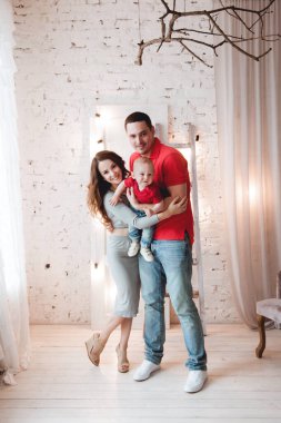 Anne ve baba, ellerinde aynı kotlu ve kırmızı tişörtlü küçük bir çocuk, parlak bir odada, perdeleri, merdivenleri ve ampulleri olan beyaz tuğladan bir duvara karşı duruyor.