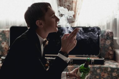 Siyah ceketli yakışıklı adam odada oturuyor ve sigara içiyor.