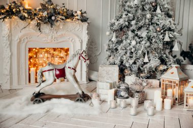 Beyaz bir odanın içinde Noel ağacı, süslü bir şömine, sallanan at ve yeni yıl hediyeleri olan bir oda.