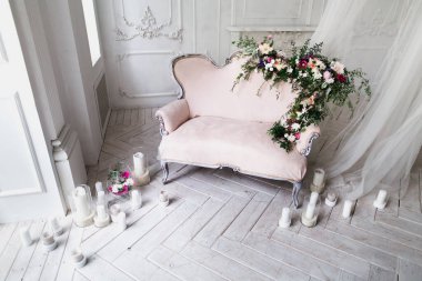 Çiçekler ve yeşilliklerle süslenmiş klasik pembe renkli kanepe, ahşap zeminde pencerenin yanındaki cam şamdanlar ve perdelerle aydınlatılmış beyaz mumlarla çevrili klasik bir odada duruyor.