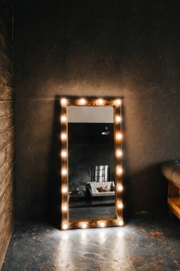 Köşe odasında ahşap ve gri beton bir duvar, yaşlanmış bir zemin, siyah cam kapılar ve ampullü elektrikli makyaj aynası ve tavan arası tarzı Oxford kanepesi.